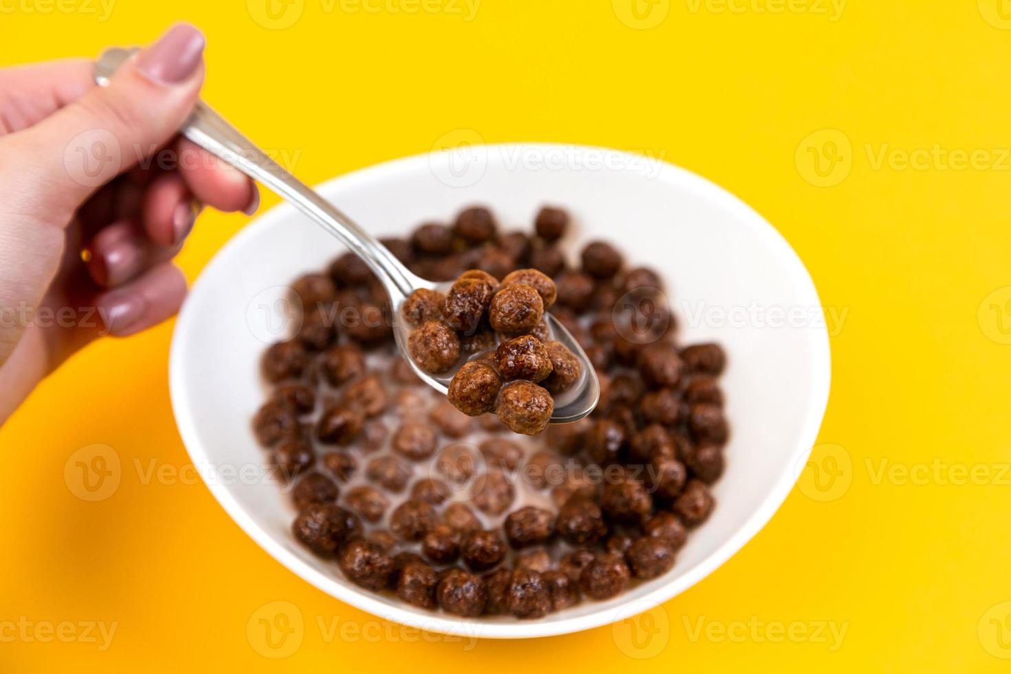 Mujer mano sosteniendo una cuchara en un tazón blanco con bolas de cereal de maíz de chocolate y leche sobre fondo amarillo foto