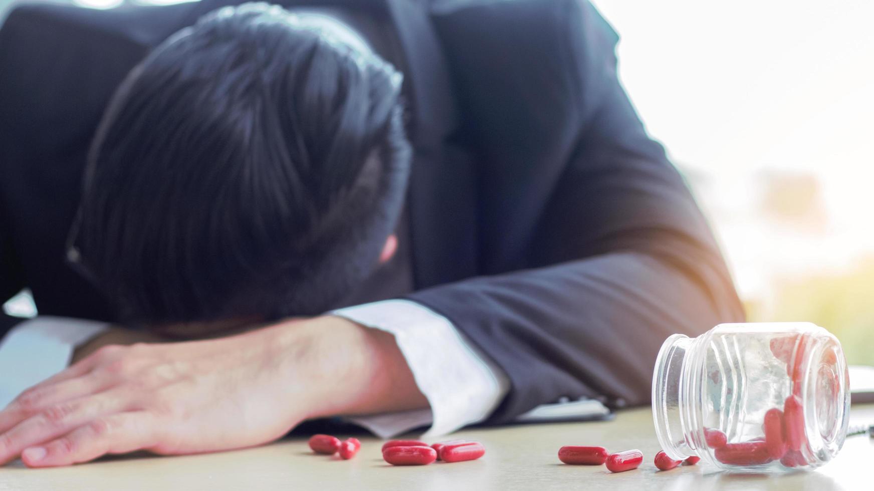 Asian businessman sleeps on his desk in fatigue with scattered painkillers on the table photo
