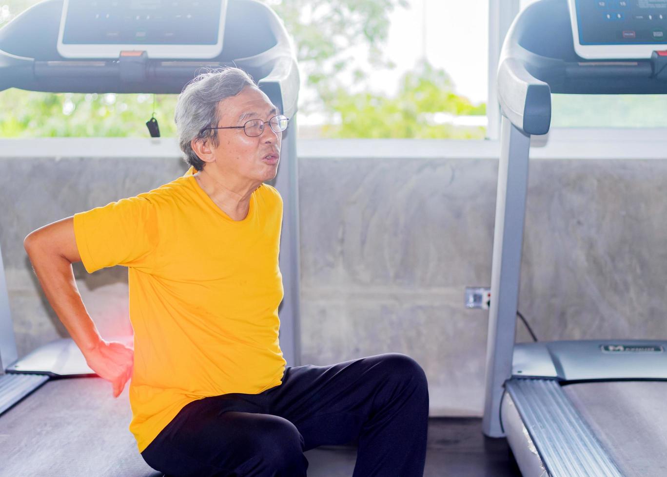 Un hombre asiático con gafas sentado en el suelo debido al dolor en la espalda por hacer ejercicio en el gimnasio foto