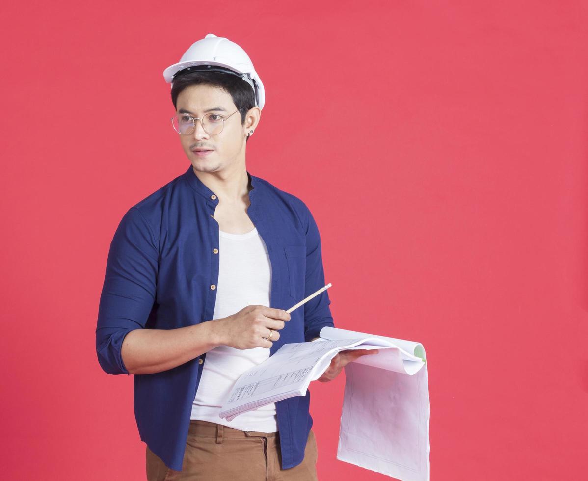 Retrato de joven arquitecto con un casco blanco sosteniendo un plan de construcción sobre un fondo rojo. foto