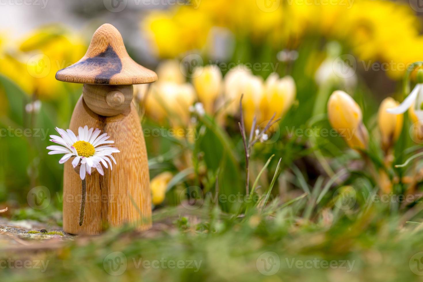 Wooden elf figure with a daisy in hand is standing in a flower bed in front of blurred flowers photo
