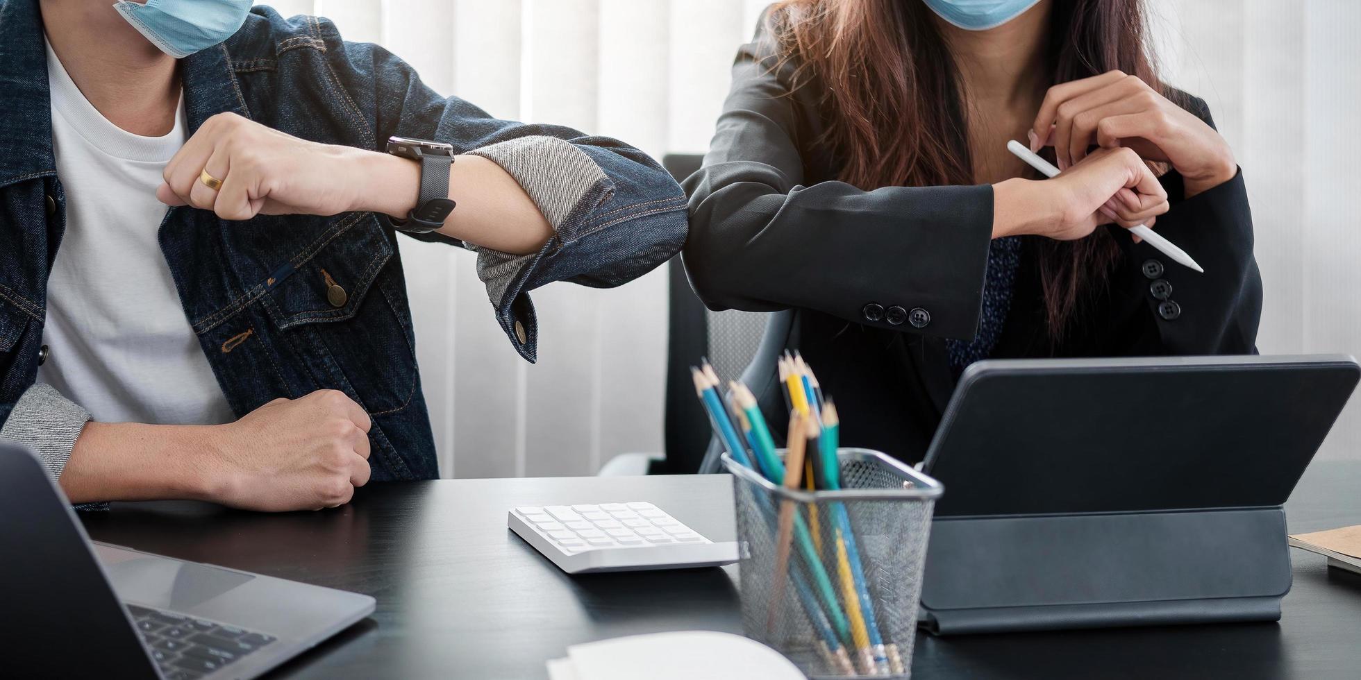 Two young diverse business colleagues wearing face protective masks bumping elbows greeting each other while working during covid 19 quarantine photo