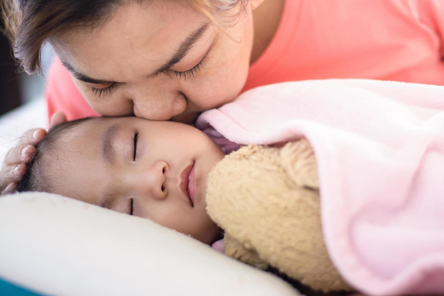 Cerrar madre asiática besando a su niña durmiendo en la cama foto