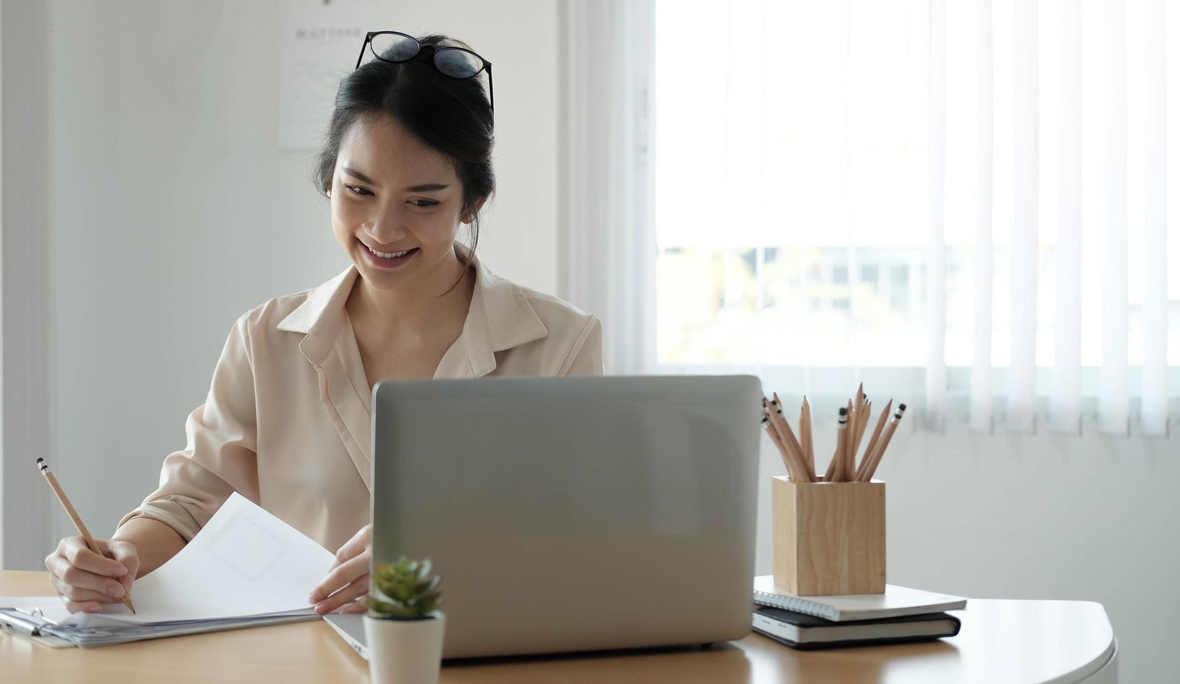 Feliz joven empresaria asiática mujer de negocios usando la computadora mirando la pantalla trabajando en internet sentarse en el escritorio de la oficina sonriente empleada profesional escribiendo correo electrónico en la computadora portátil en el lugar de trabajo foto