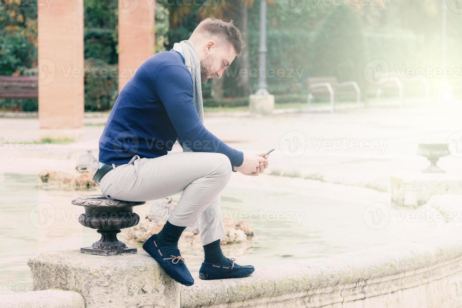young man sitting in the fountain of a park looking at his cell phone photo