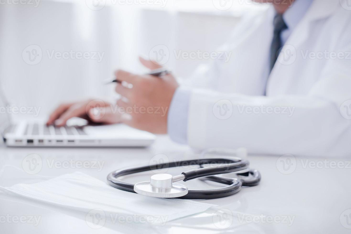 Doctor working with laptop computer and writing on paperwork Hospital background Healthcare and medical concept Focus on stethoscope photo