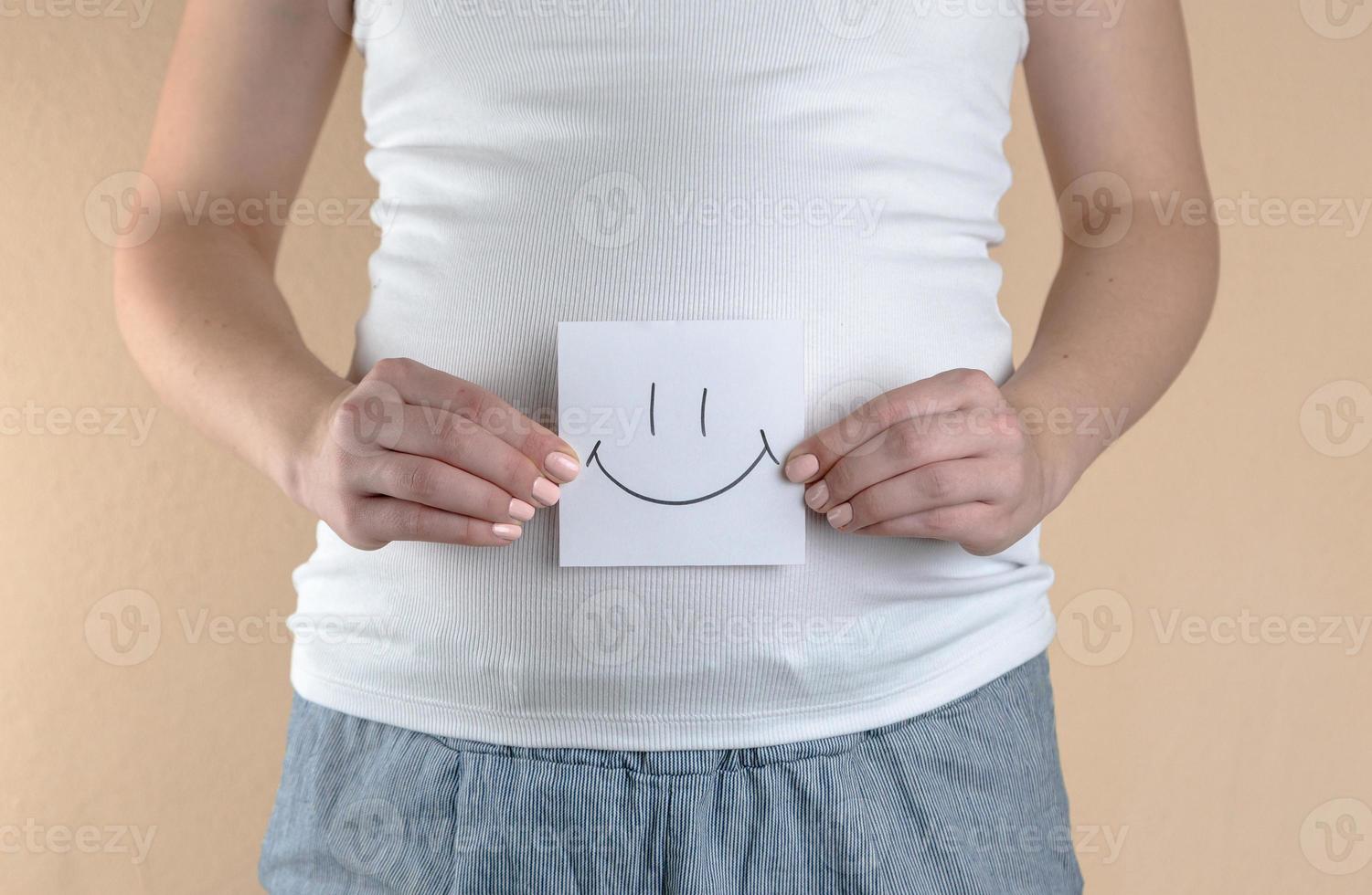 A close up view of the belly of a pregnant woman that is holding a piece of paper with a smiley face photo