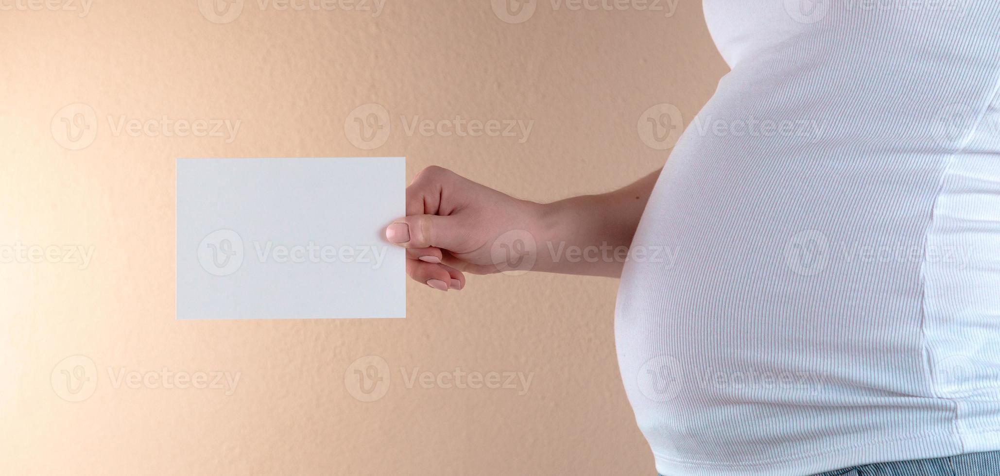 Una vista de cerca del vientre de una mujer embarazada que sostiene una hoja de papel vacía foto