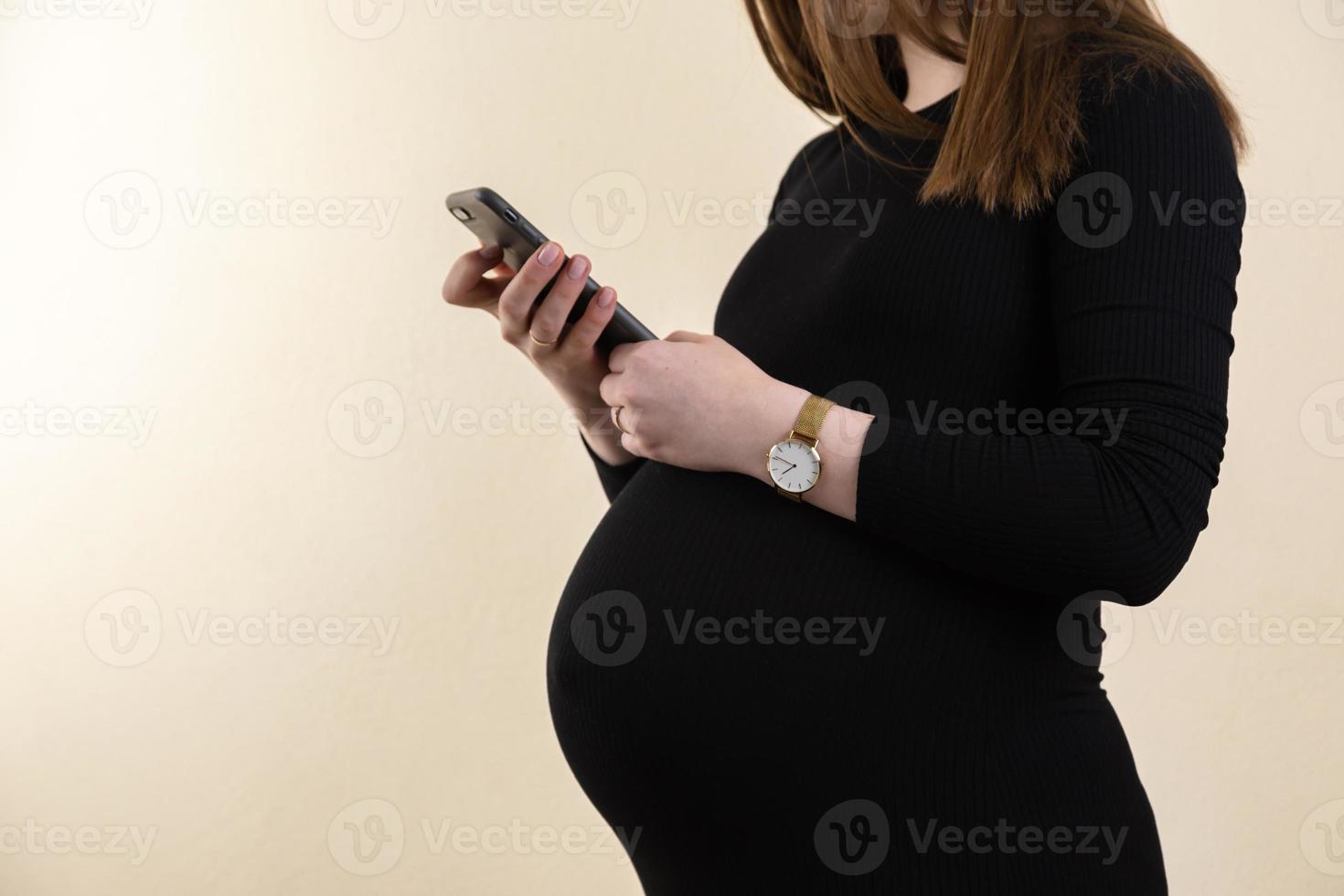 Joven embarazada con un vestido negro sostiene su vientre y mira su teléfono foto
