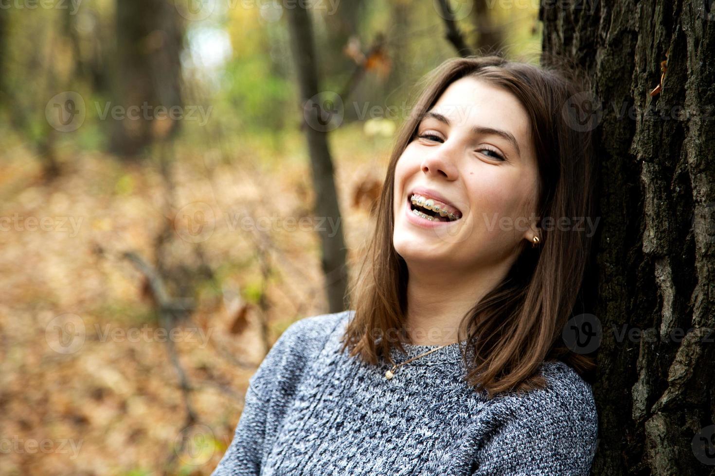 Young beautiful girl with braces on her teeth in a gray sweater sits in the autumn forest photo