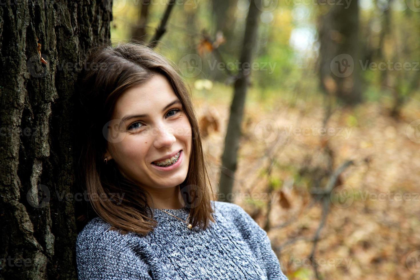 Young beautiful girl with braces on her teeth in a gray sweater sits in the autumn forest photo