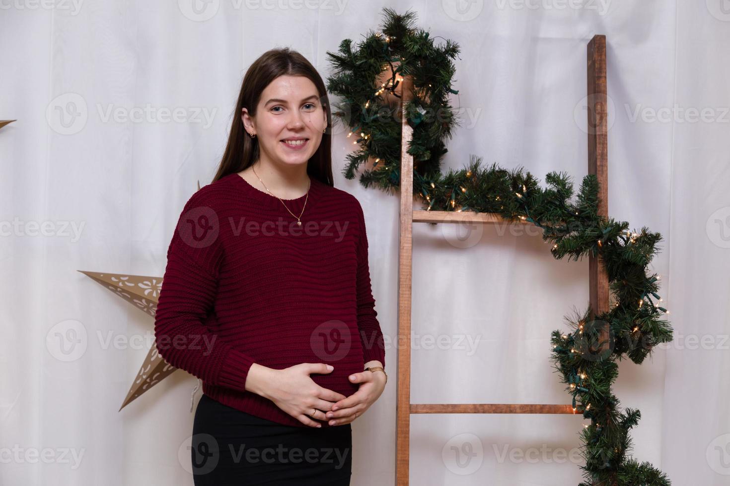 Hermosa niña embarazada sonriente de pie en el fondo de la decoración navideña foto