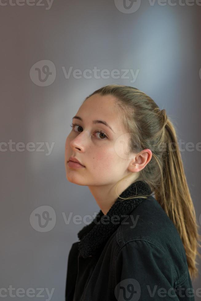 Tiro de tres cuartos o retrato de semi perfil de una bonita adolescente con el pelo de cola de caballo posando mirando a la cámara sobre un fondo gris foto