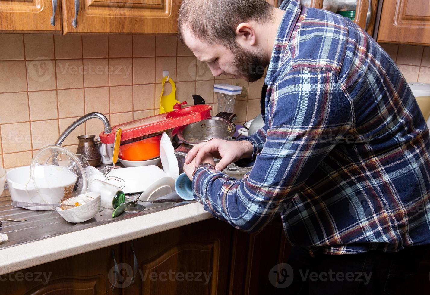 A young bearded guy looks at his watch and is shocked by the amount of dirty dishes lying in the kitchen sink photo