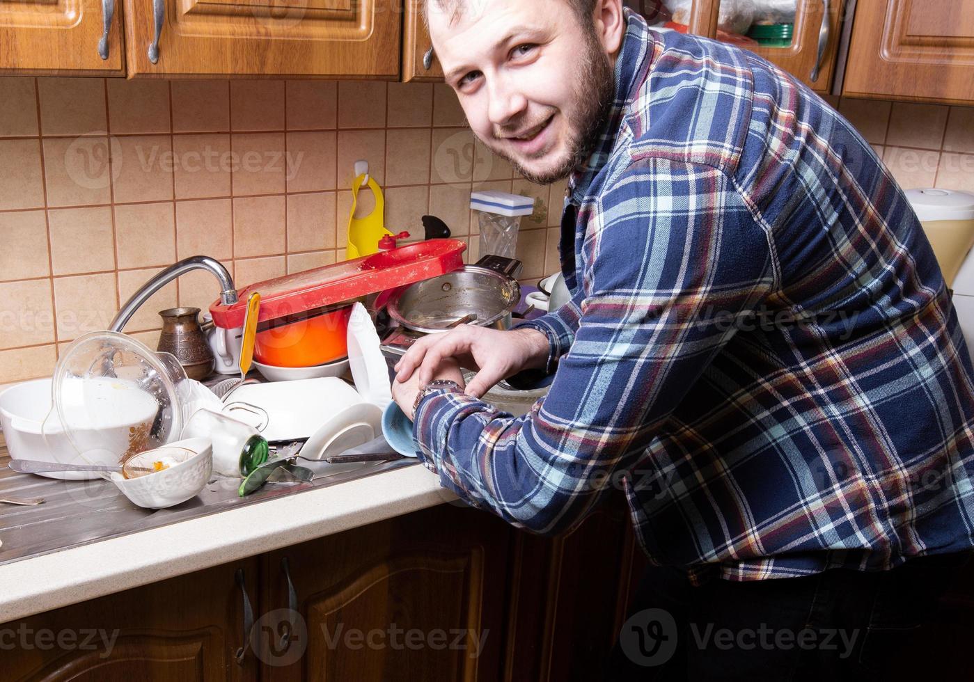 A young bearded guy looks at his watch and is happy by the amount of dirty dishes lying in the kitchen sink photo