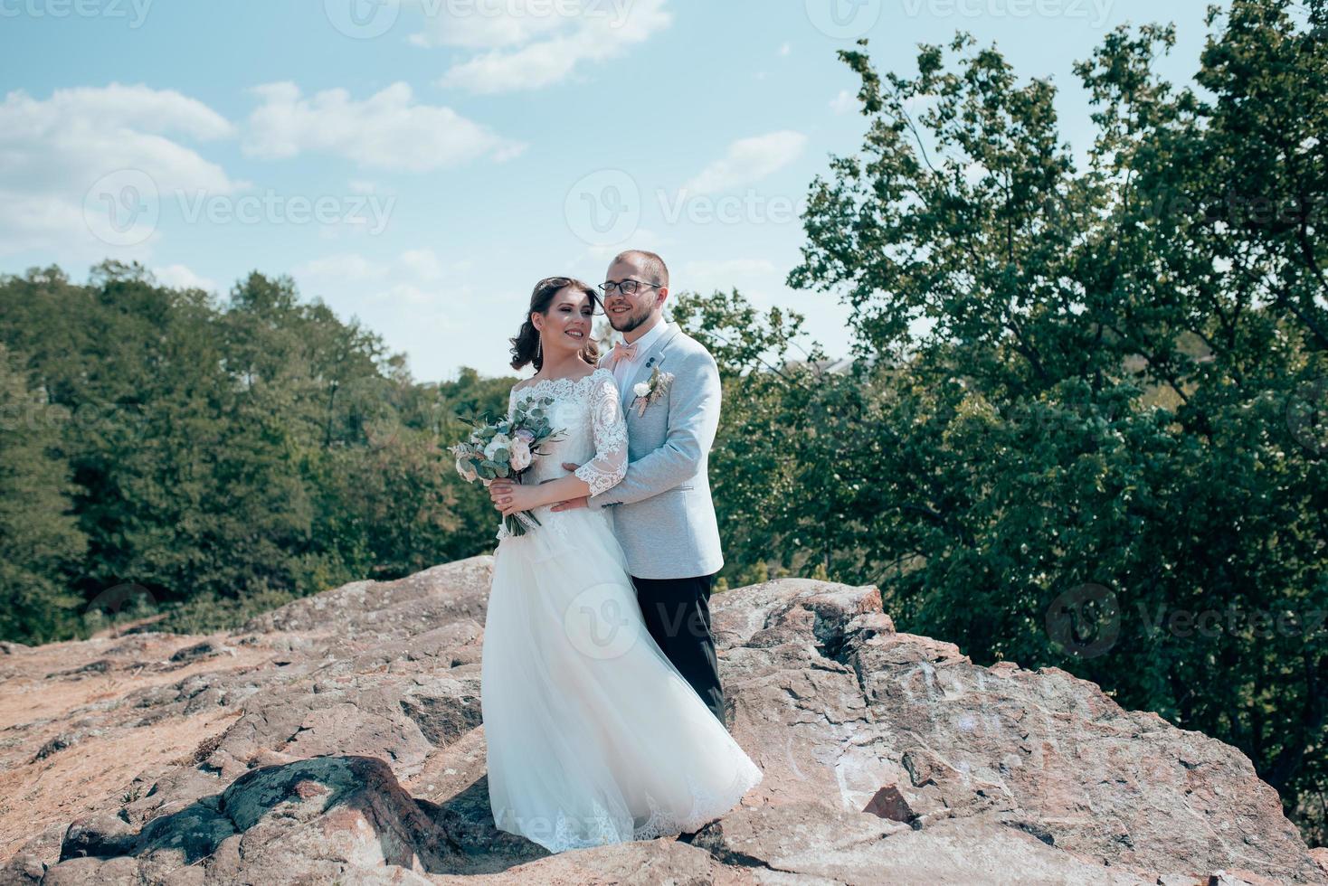 Foto de boda de un novio barbudo con gafas en una chaqueta gris y una novia en una roca