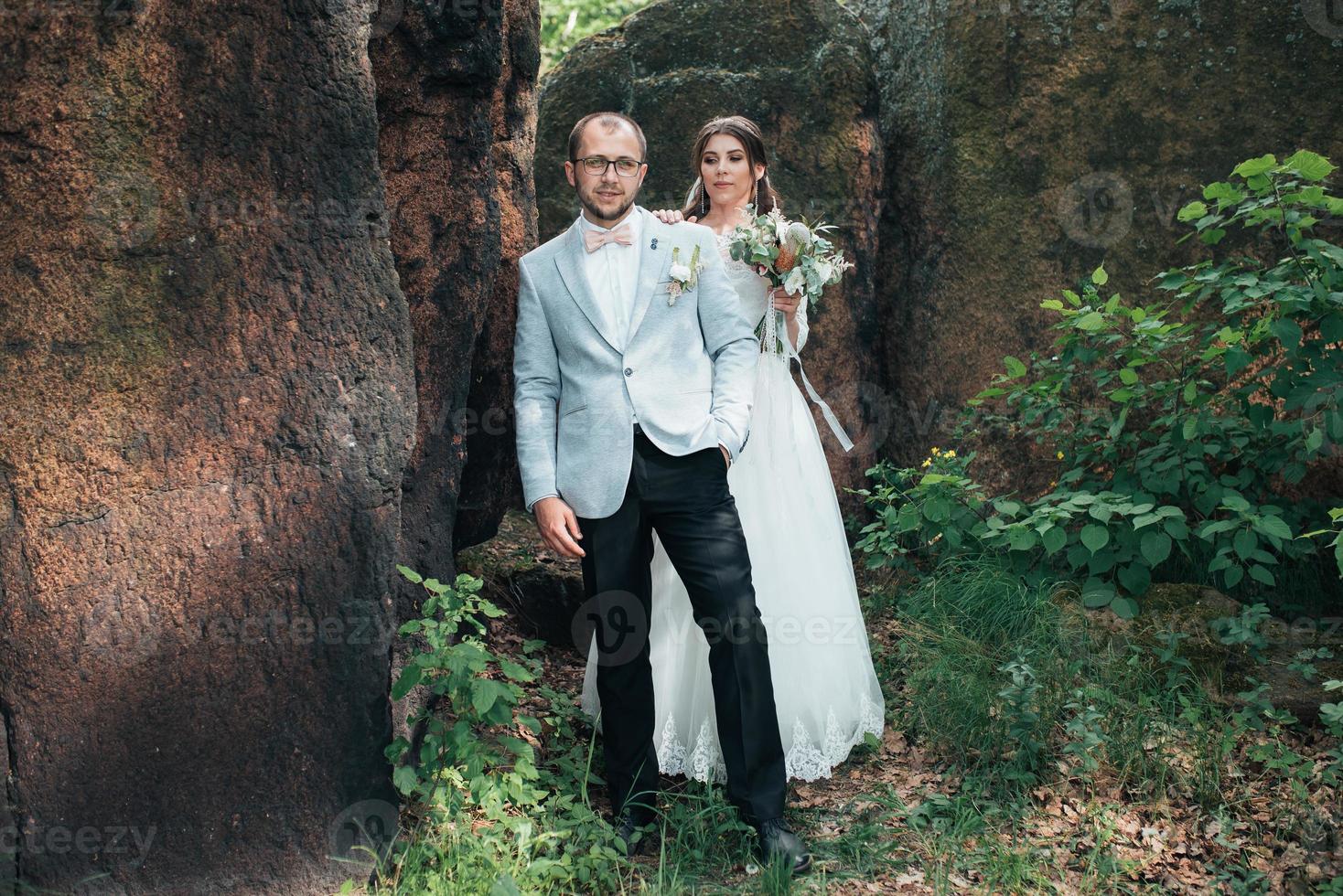 La novia y el novio en el día de la boda abrazándose de pie cerca de una roca o una piedra grande foto