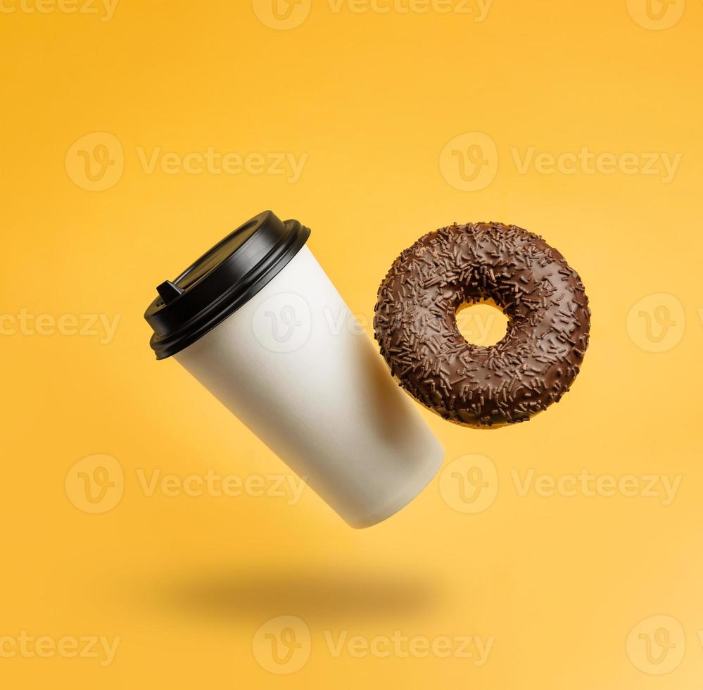 A glass with coffee and a chocolate donut levitating against a yellow background with a copy space photo