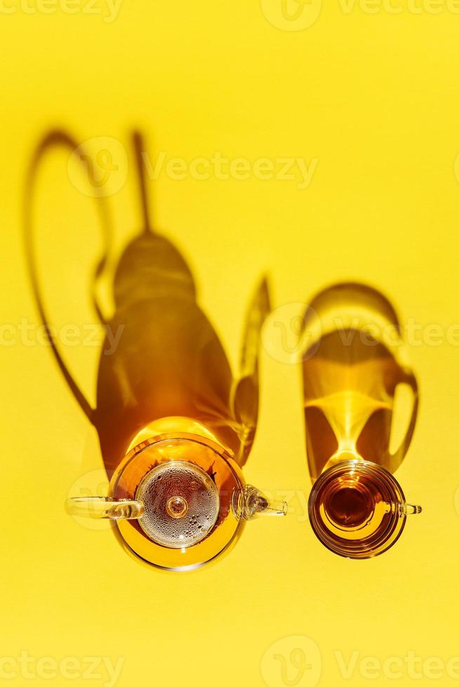 A glass teapot and a cup with tea shot from above show off long shadows on a bright yellow background photo