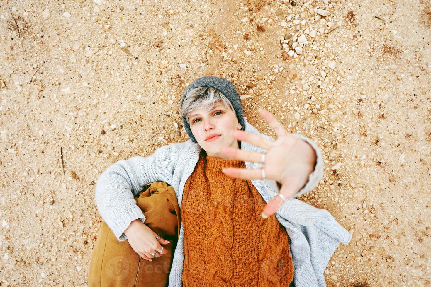 An adventurer young caucasian woman lying on grit ground beside a backpack wearing wool sweater and cap with her hand open and the orange as principal color photo