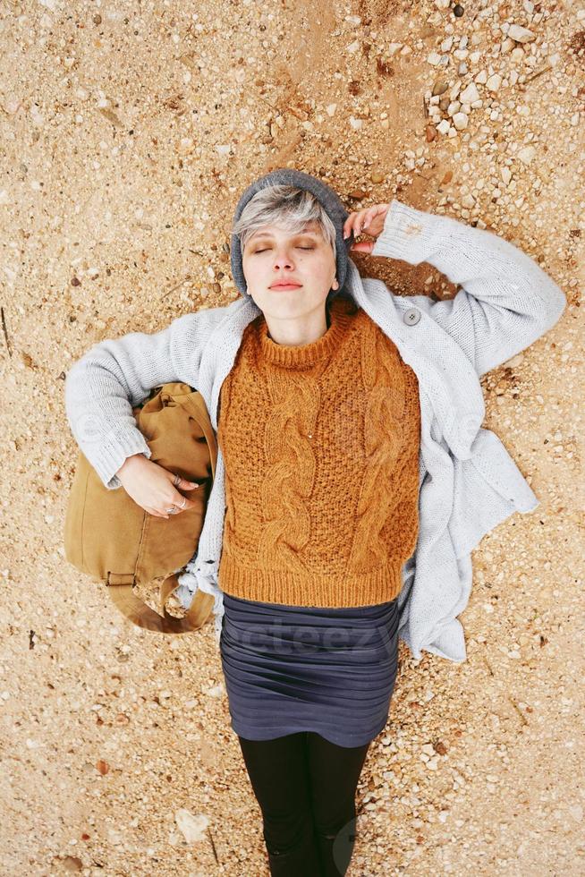 Un aventurero joven mujer caucásica tumbado en el suelo junto a una mochila vistiendo un suéter de lana y una gorra con los ojos cerrados y el naranja como color principal foto