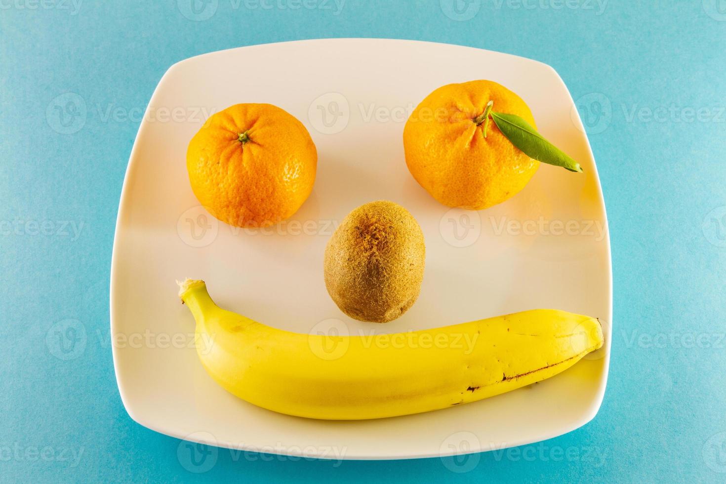 Tangerines kiwi and one banana on a white plate as smiling human face photo