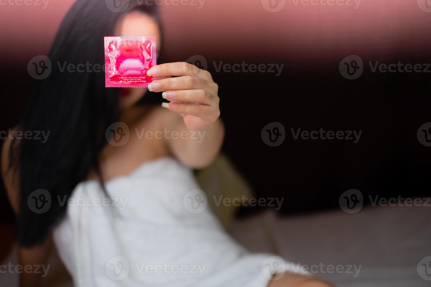 centrarse en el condón rojo en la mano el primer plano en la hermosa mujer con una toalla blanca yace en la cama foto