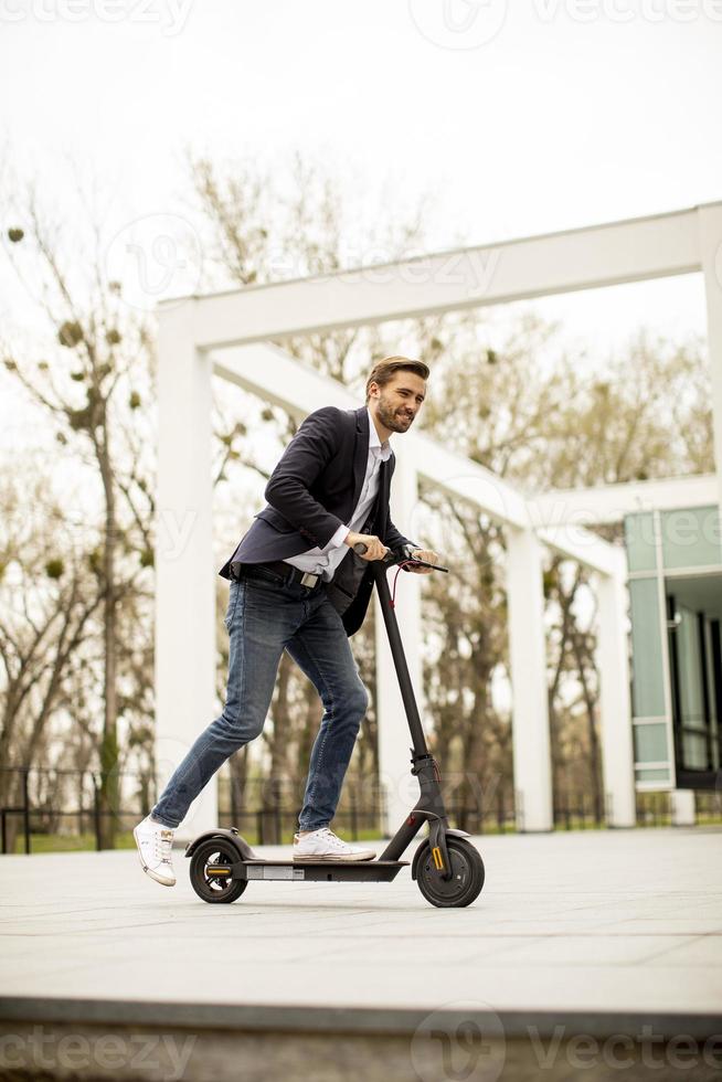 Hombre de negocios joven en ropa casual montando un scooter eléctrico por un edificio de oficinas en una reunión de negocios foto