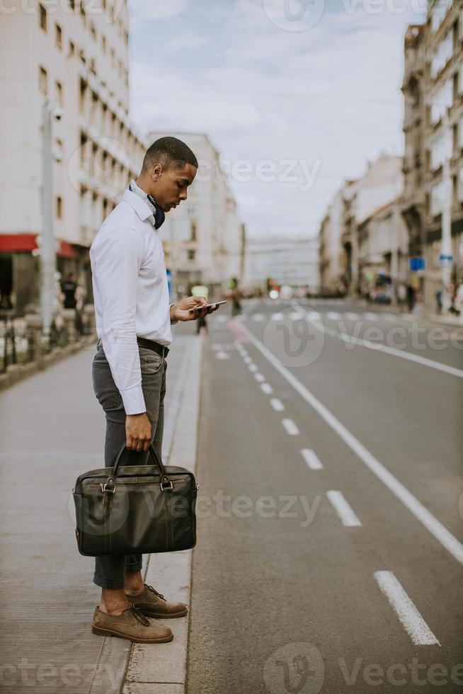 Joven empresario afroamericano mediante un teléfono móvil mientras espera un taxi en una calle foto