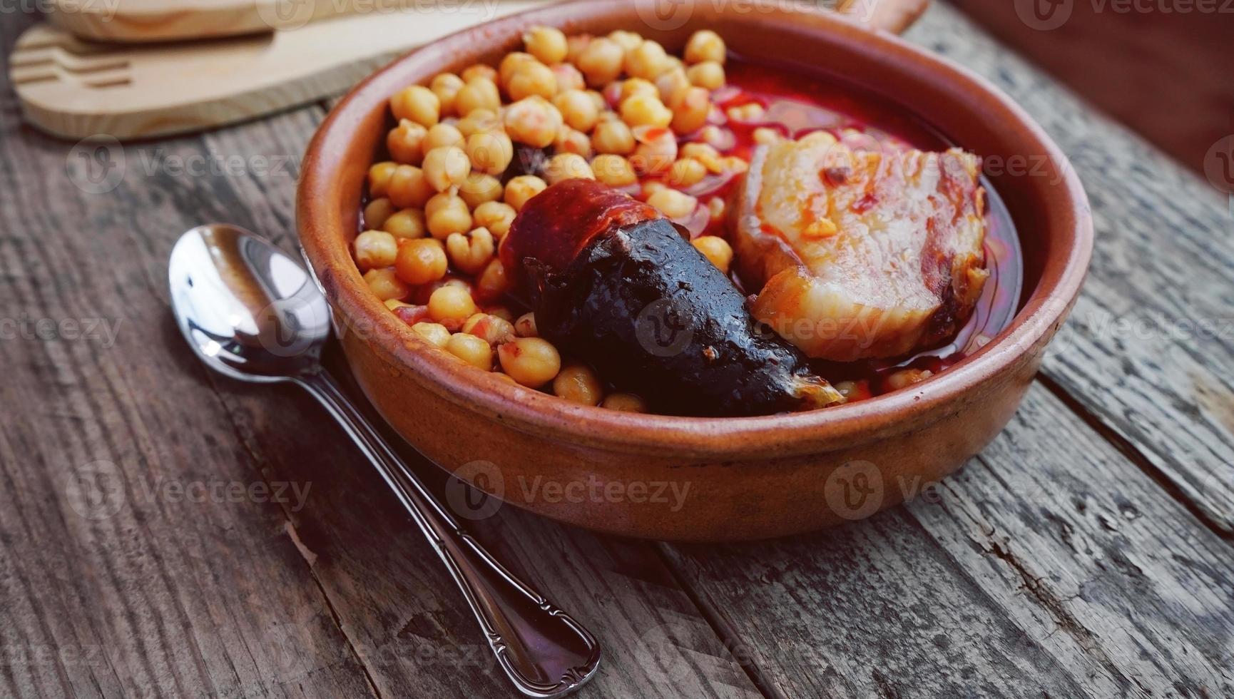 garbanzos, salchicha y tocino en una olla de cocción lenta con una cuchara de madera y un tenedor y una pequeña cuchara de metal foto