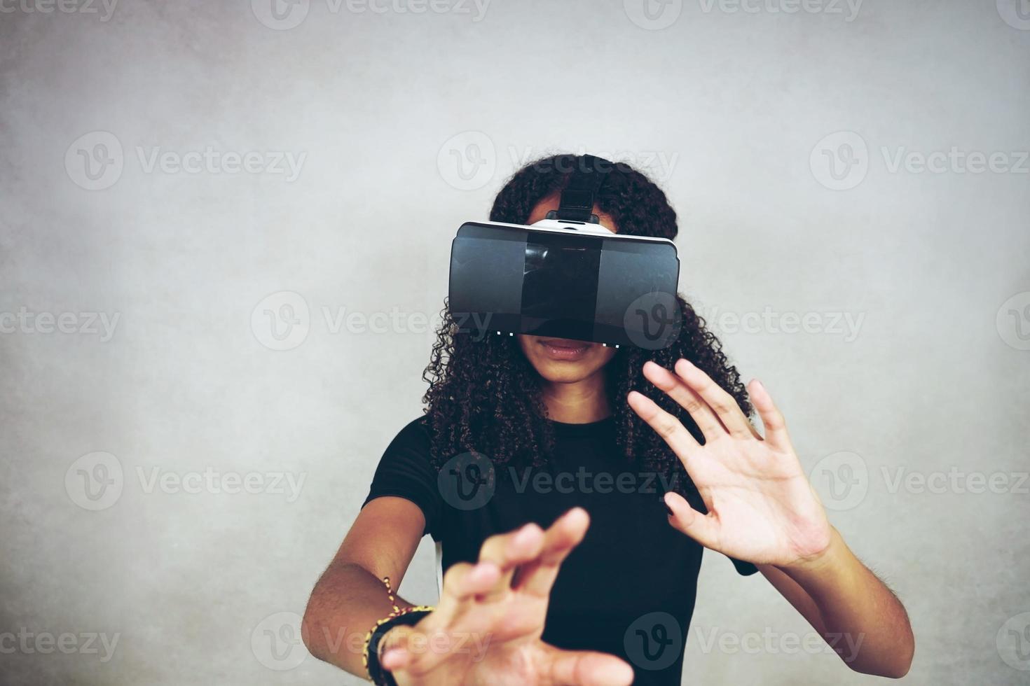 Una hermosa joven negra con cabello afro rizado usa auriculares de realidad virtual vr y juega videojuegos en estudio con fondo gris foto