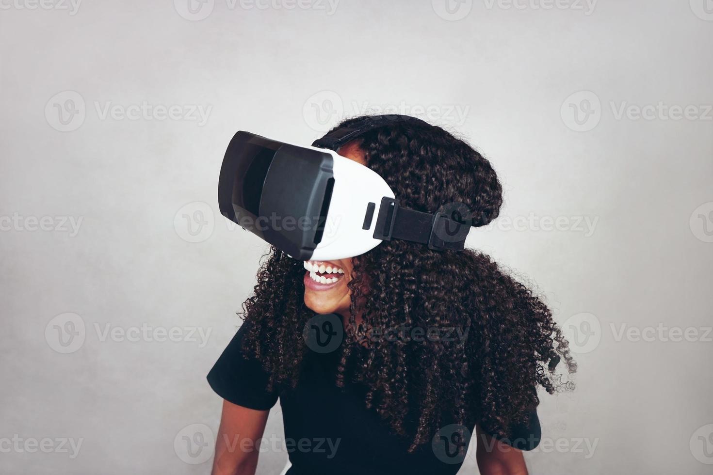 Una hermosa joven negra con cabello afro rizado usa auriculares de realidad virtual vr y juega videojuegos mientras sonríe en el estudio con fondo gris foto