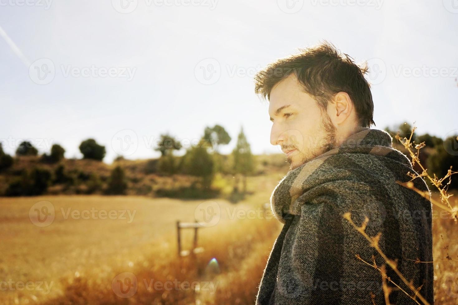 Retrato lateral de un joven disfrutando en la calma del sol de otoño de la mañana en un camino de un campo amarillo con la luz de fondo del cielo azul foto