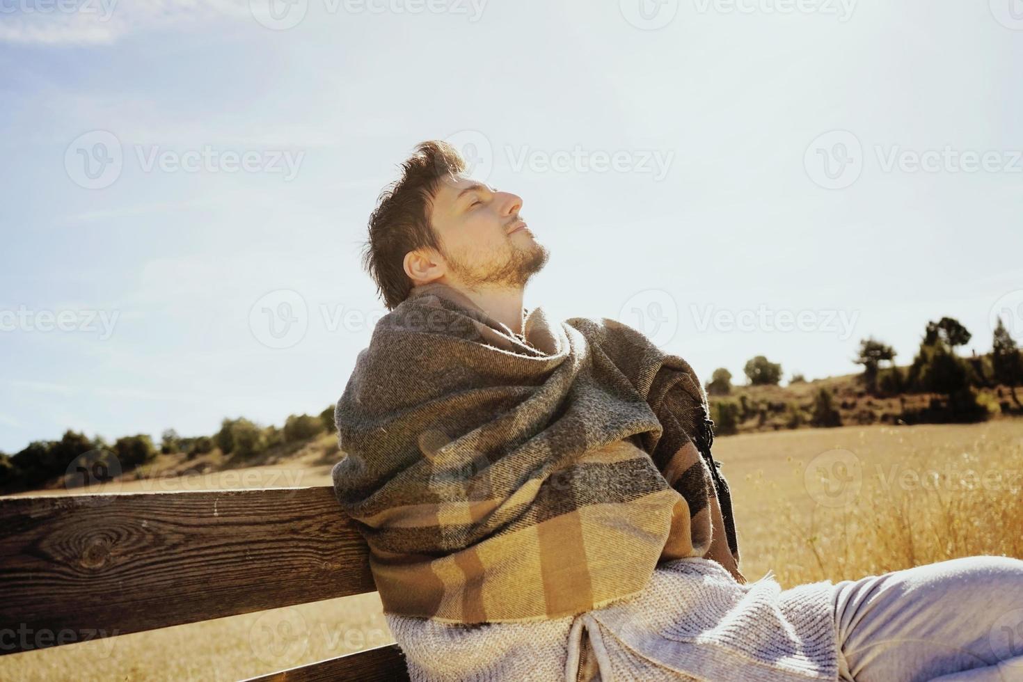 Cara lateral de un hombre joven con los ojos cerrados disfrutando en calma del sol de otoño de la mañana en un campo amarillo con la luz de fondo del cielo azul foto