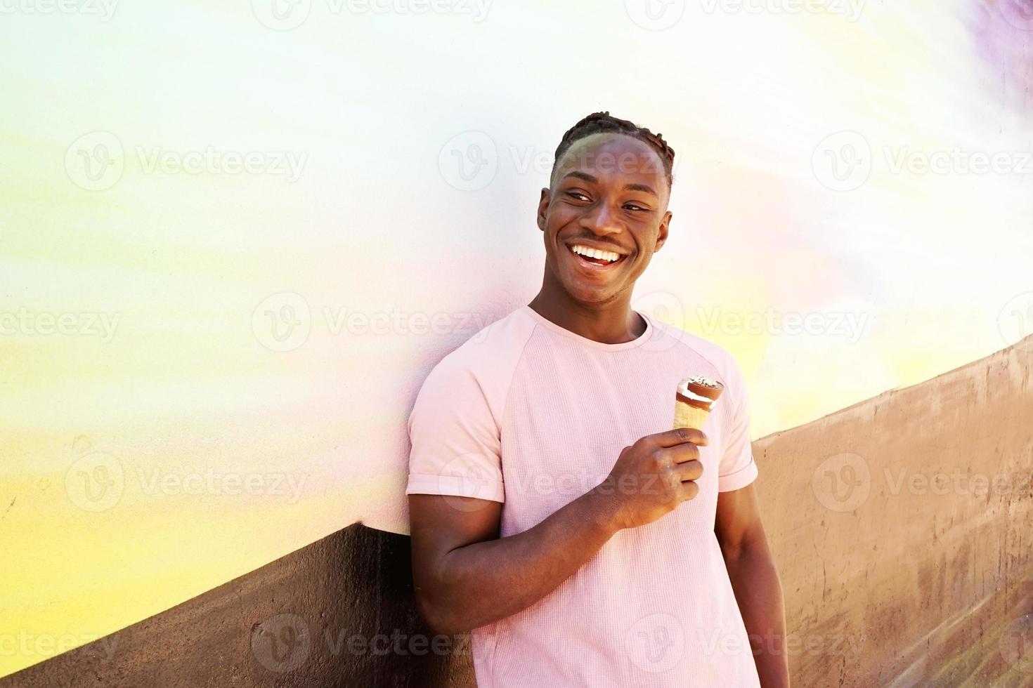 Joven guapo negro viste una camisa rosa sostiene y come un cono de helado en verano en una pared pintada como un amanecer o un día soleado foto