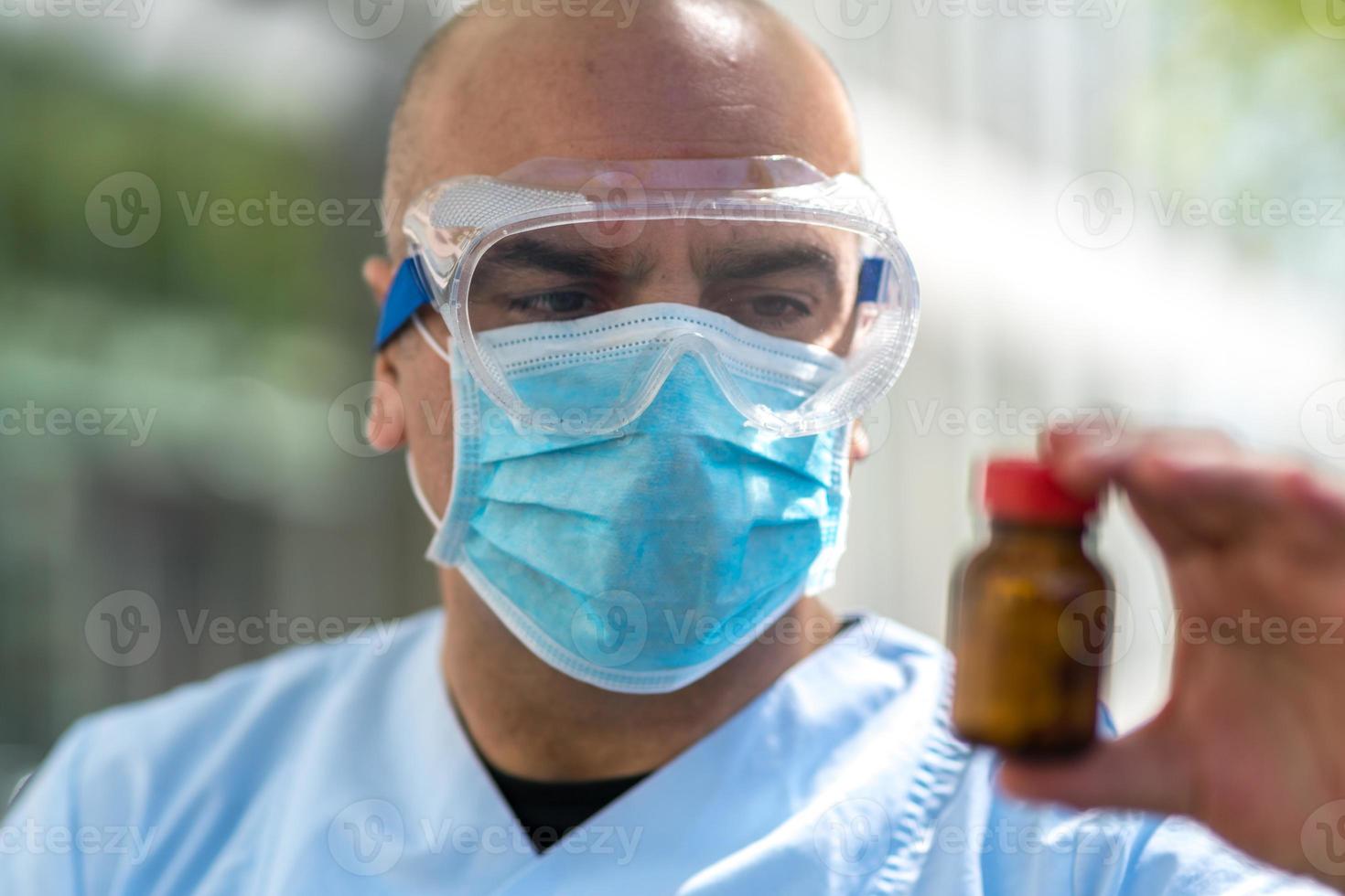 Out of focused doctor wearing lab coat, surgical mask and medical safety goggles with a bottle of liquid antibiotics photo