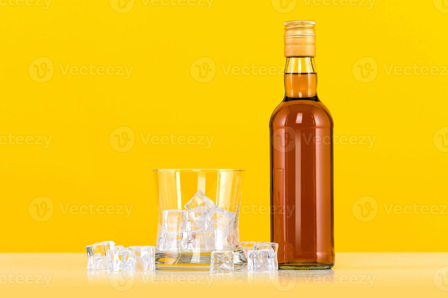 Glass of whiskey with ice cubes and bottle on yellow background photo