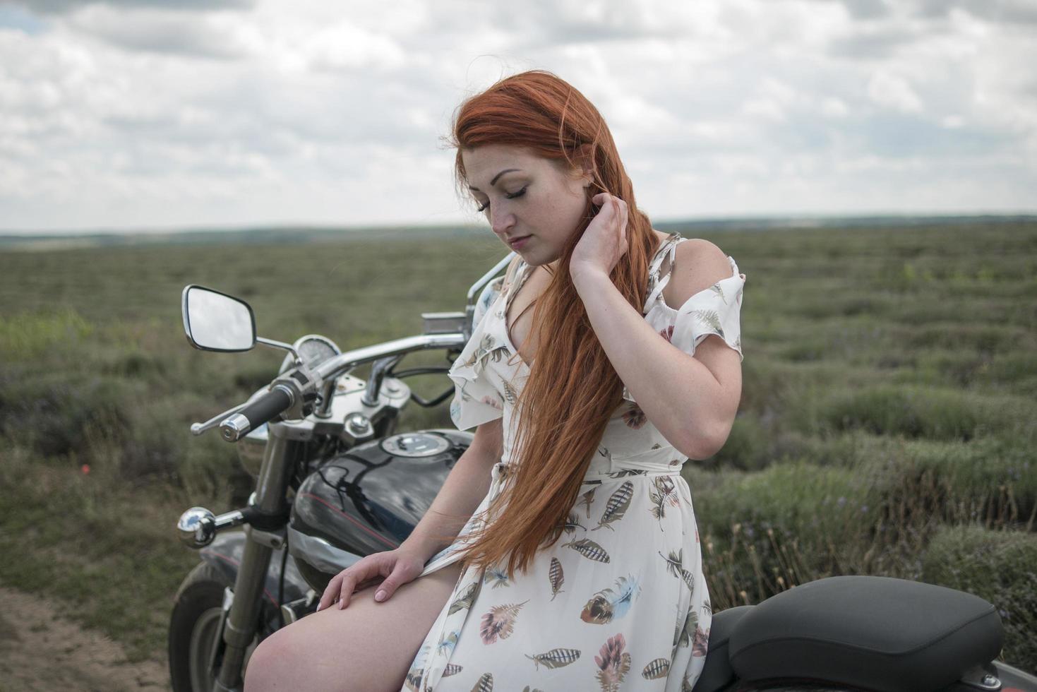 Chica pelirroja con un vestido blanco y botas junto con un campo de lavanda en motocicleta foto