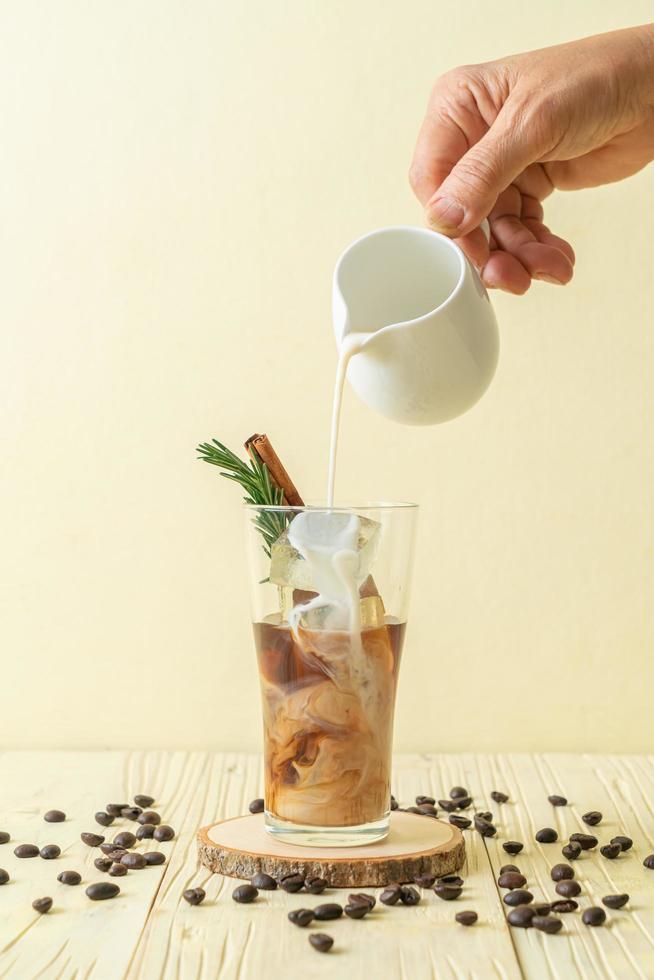 Pouring milk in black coffee glass with ice cubes, cinnamon and rosemary on wood background photo
