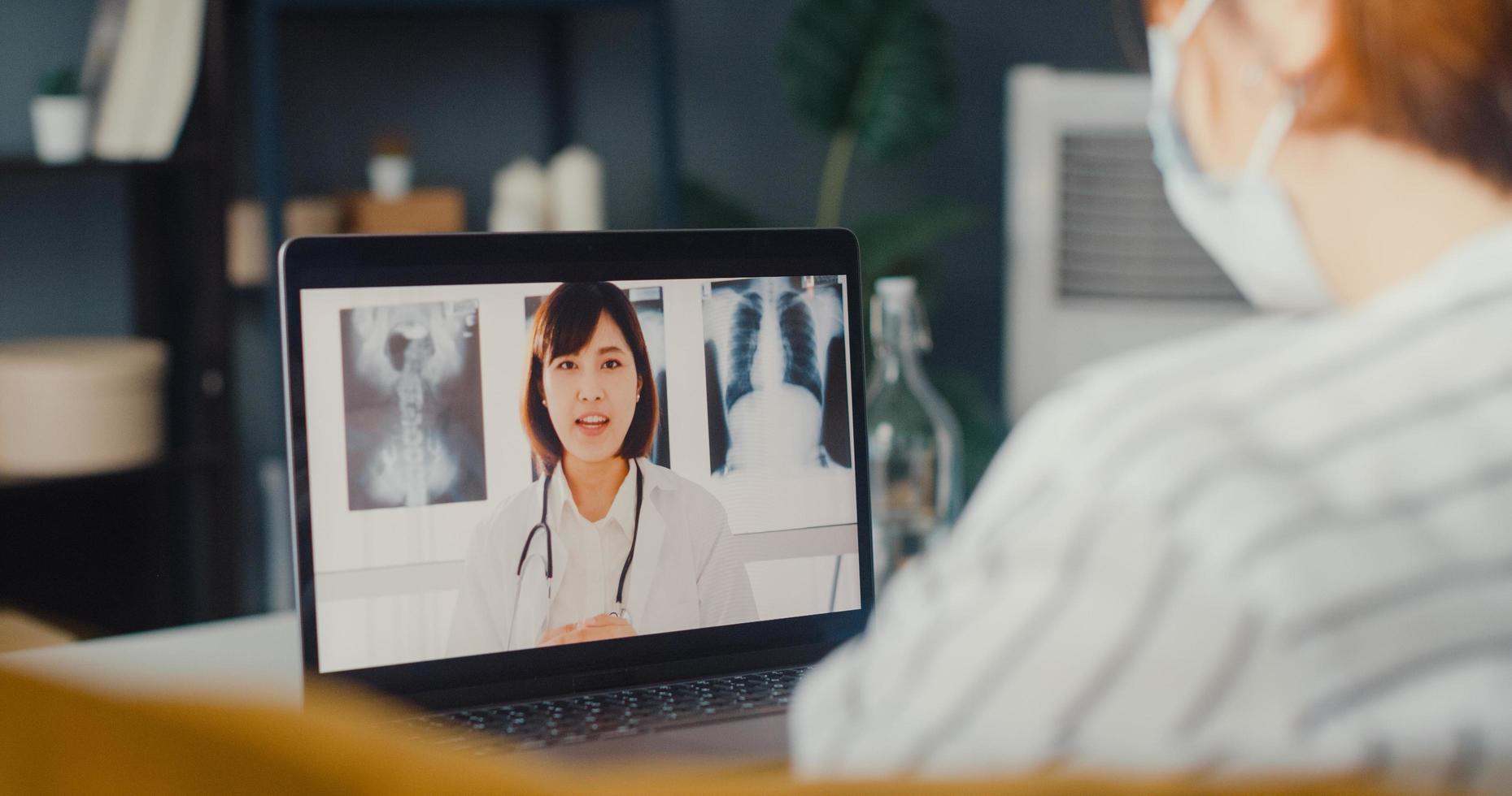 Una joven asiática usa una mascarilla protectora usando una computadora portátil, habla sobre la enfermedad en una videollamada con un médico senior, consulta en línea en la sala de estar de la casa foto