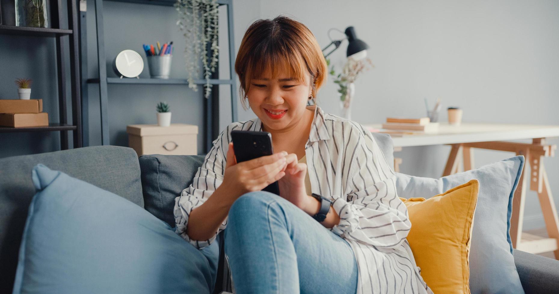 Señora joven de Asia con mensaje de texto de teléfono inteligente o comprobar las redes sociales en el sofá en la sala de estar en casa foto