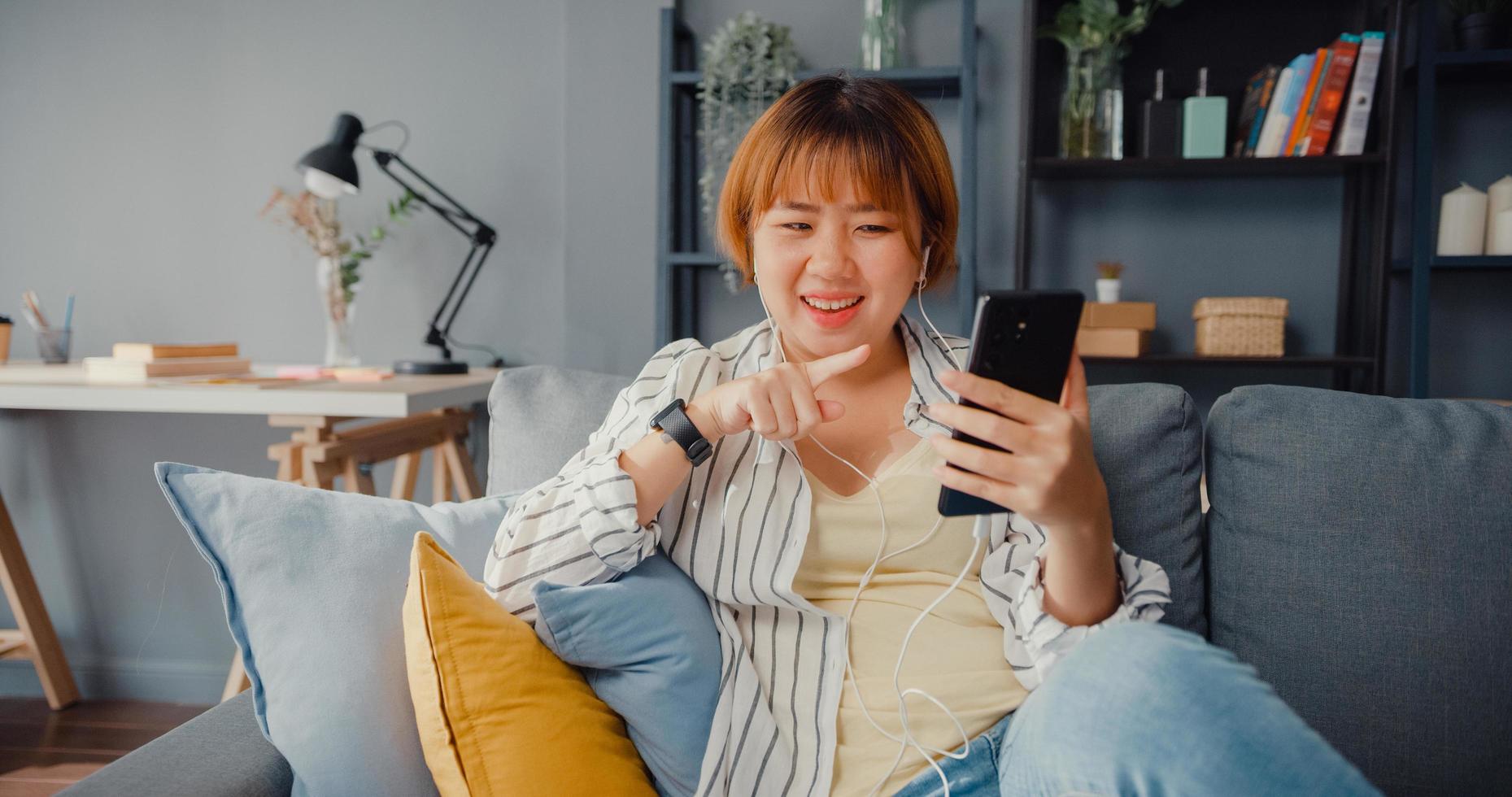 Señora joven de Asia mediante videollamada de teléfono inteligente hablar con la familia en el sofá en el salón de la casa foto