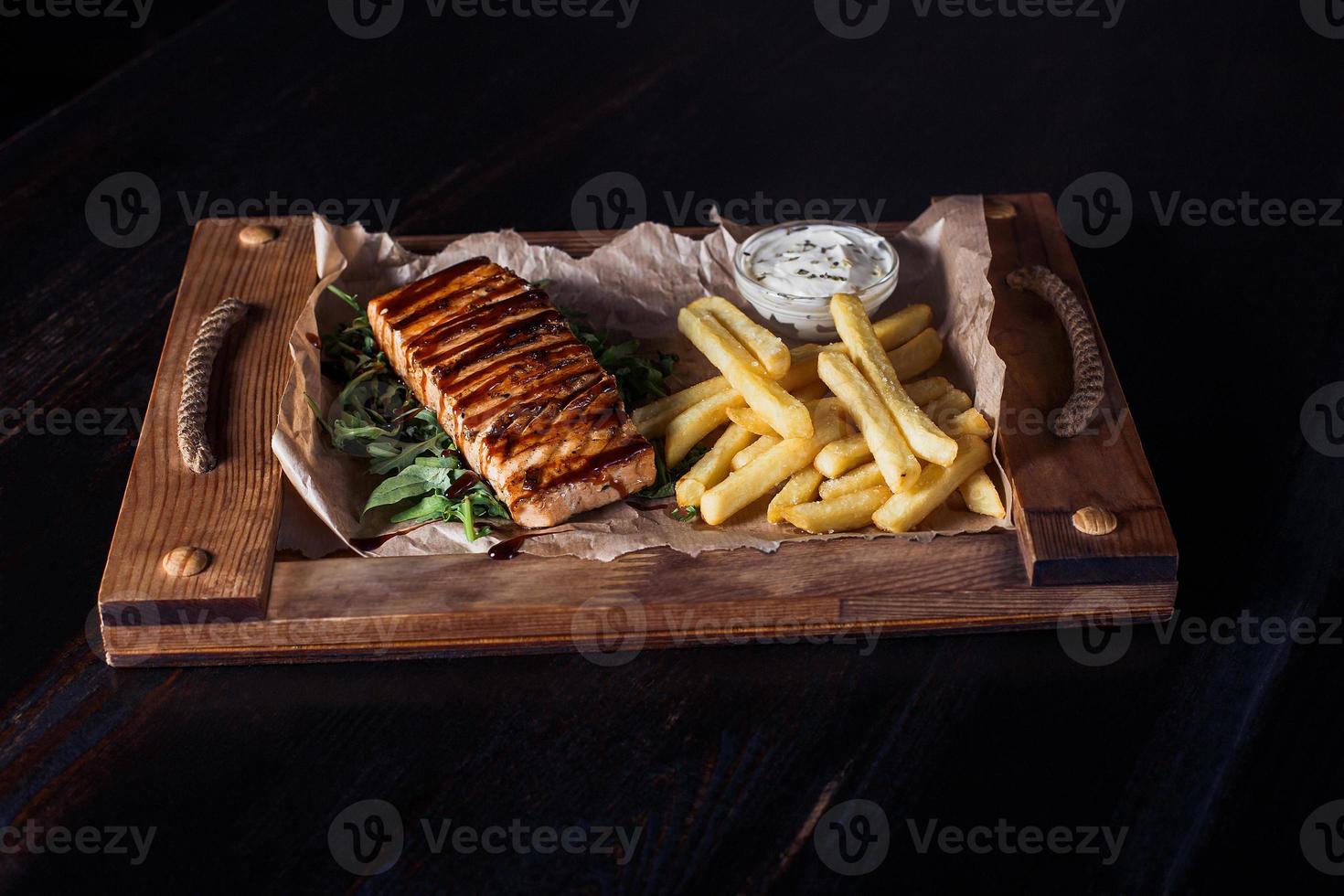 Filete de salmón con papas fritas en una bandeja de madera, hermosa porción, fondo oscuro foto