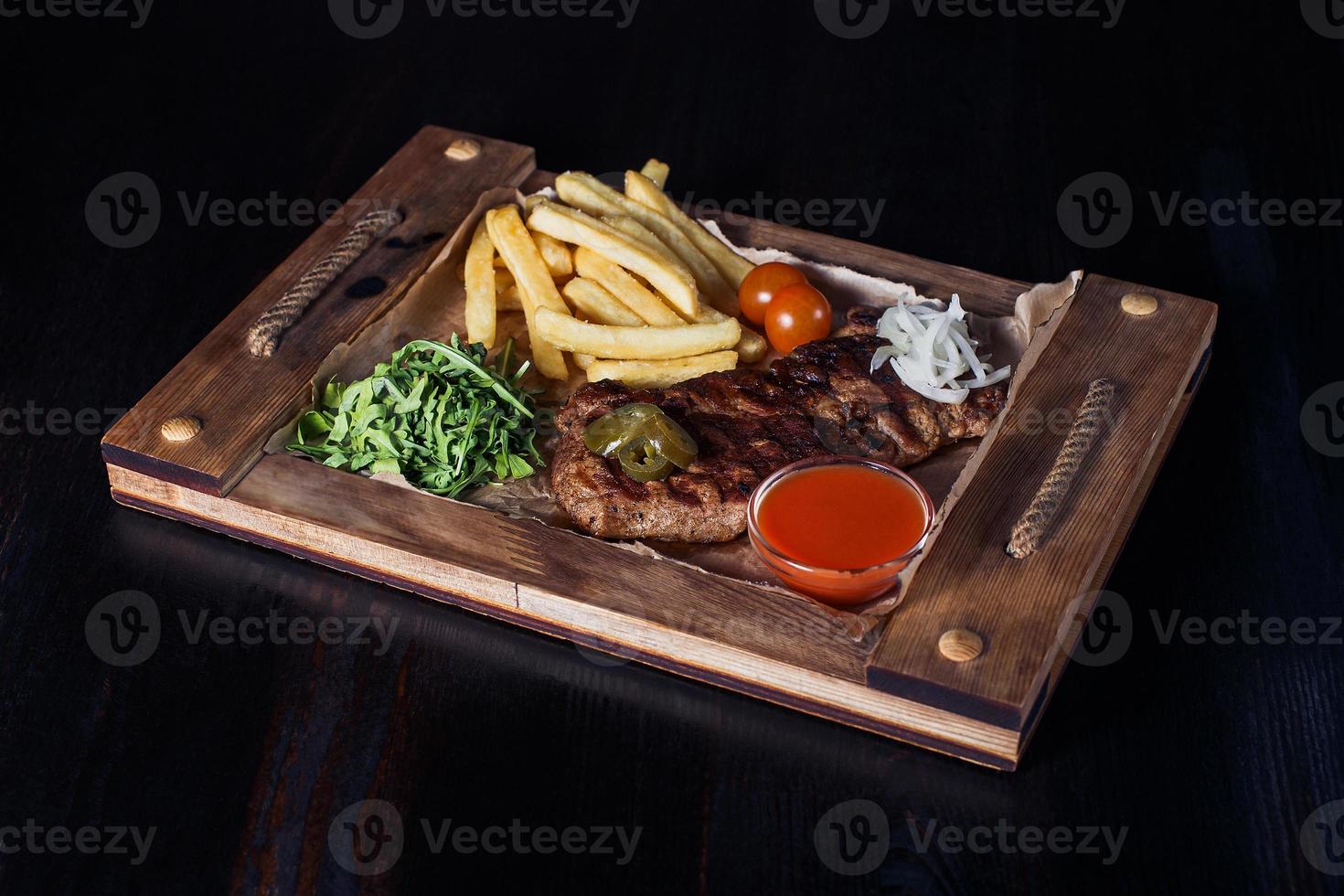 Filete de ternera con papas fritas en una bandeja de madera, hermosa porción, fondo oscuro foto