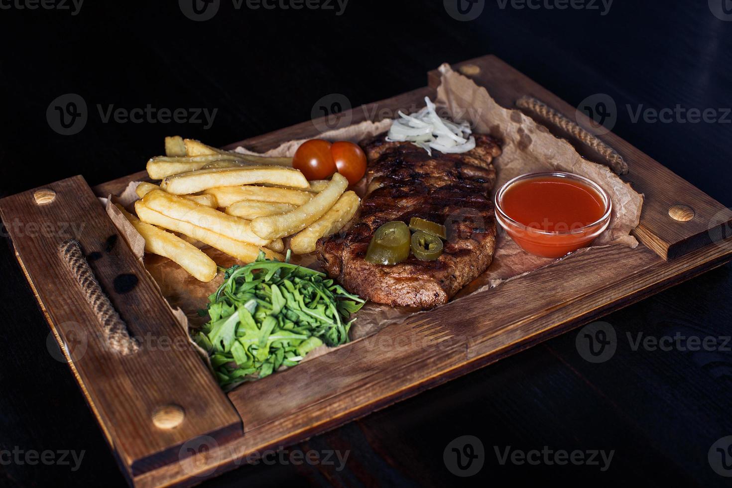 Filete de ternera con papas fritas en una bandeja de madera, hermosa porción, fondo oscuro foto
