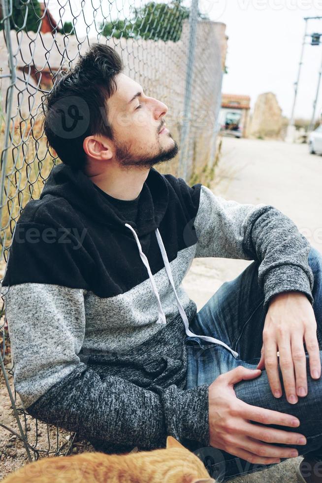 Un joven atractivo en calma sentado en el suelo y una valla al aire libre con los ojos cerrados y la cabeza hacia arriba foto