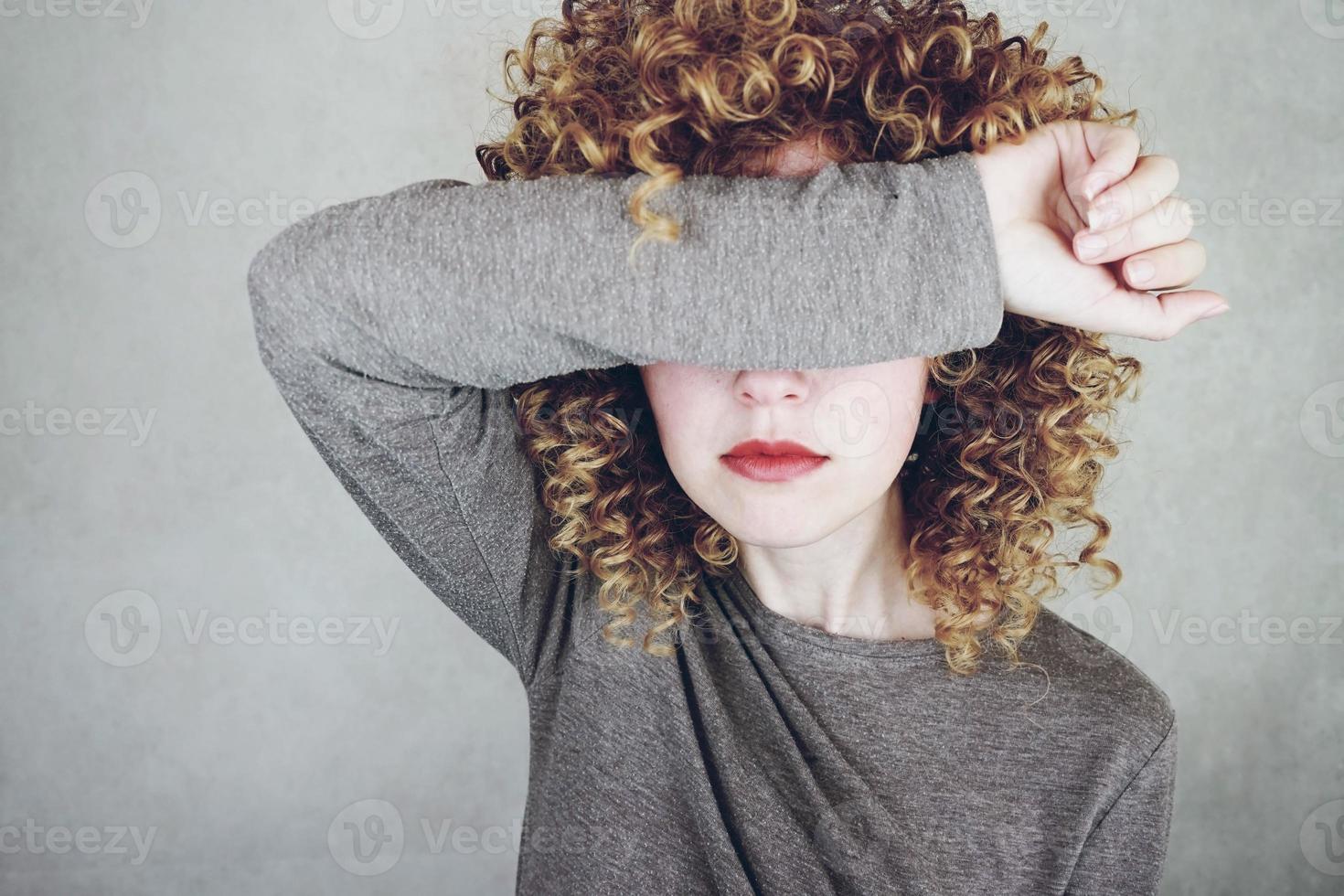 Primer plano de una hermosa y joven mujer con cabello rubio rizado cubre su rostro con su brazo, parece enojada o cansada foto