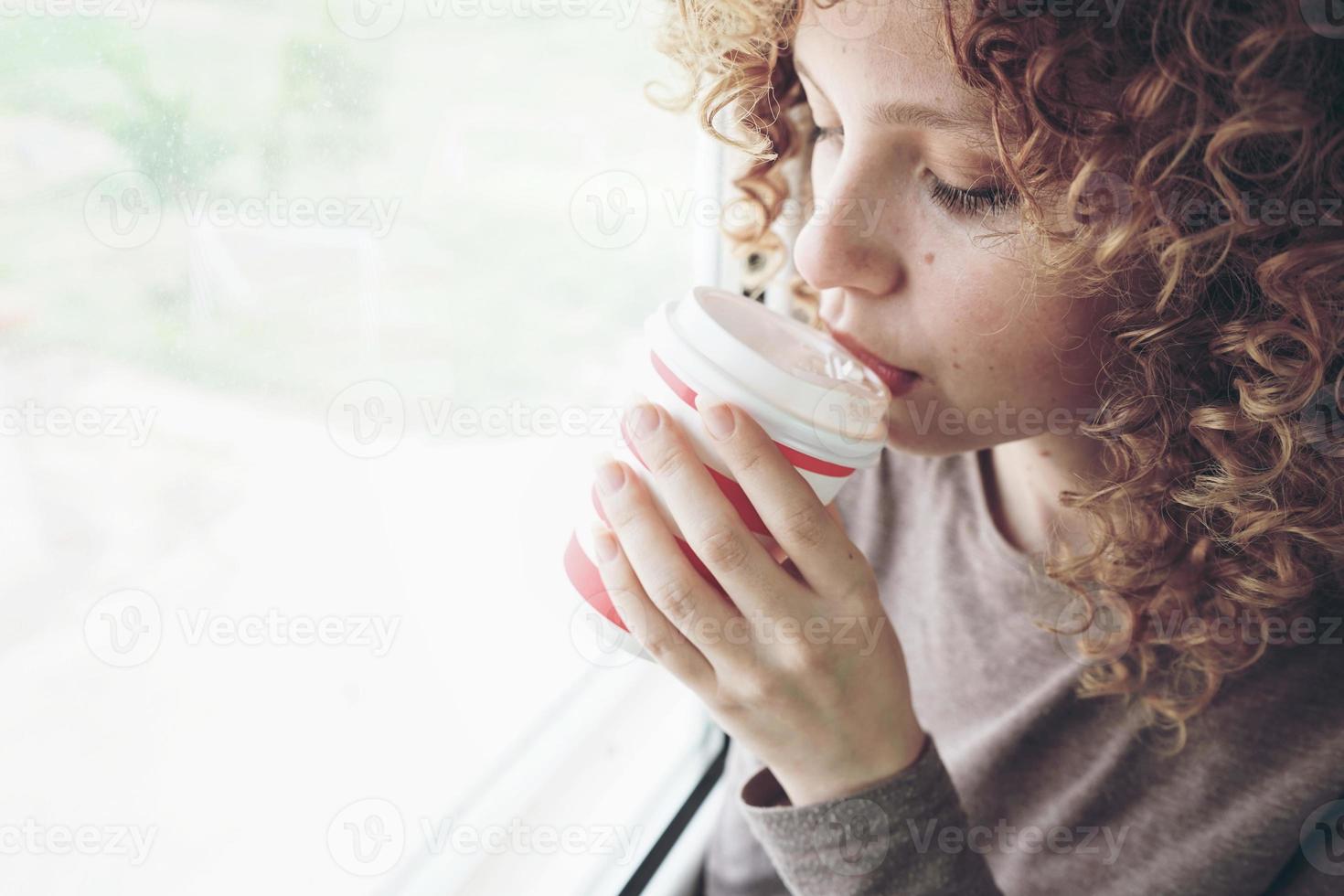 Closeup portrait of a beautiful and young woman with blue eyes and curly blonde hair drinks coffee or tea while she is in a journey photo