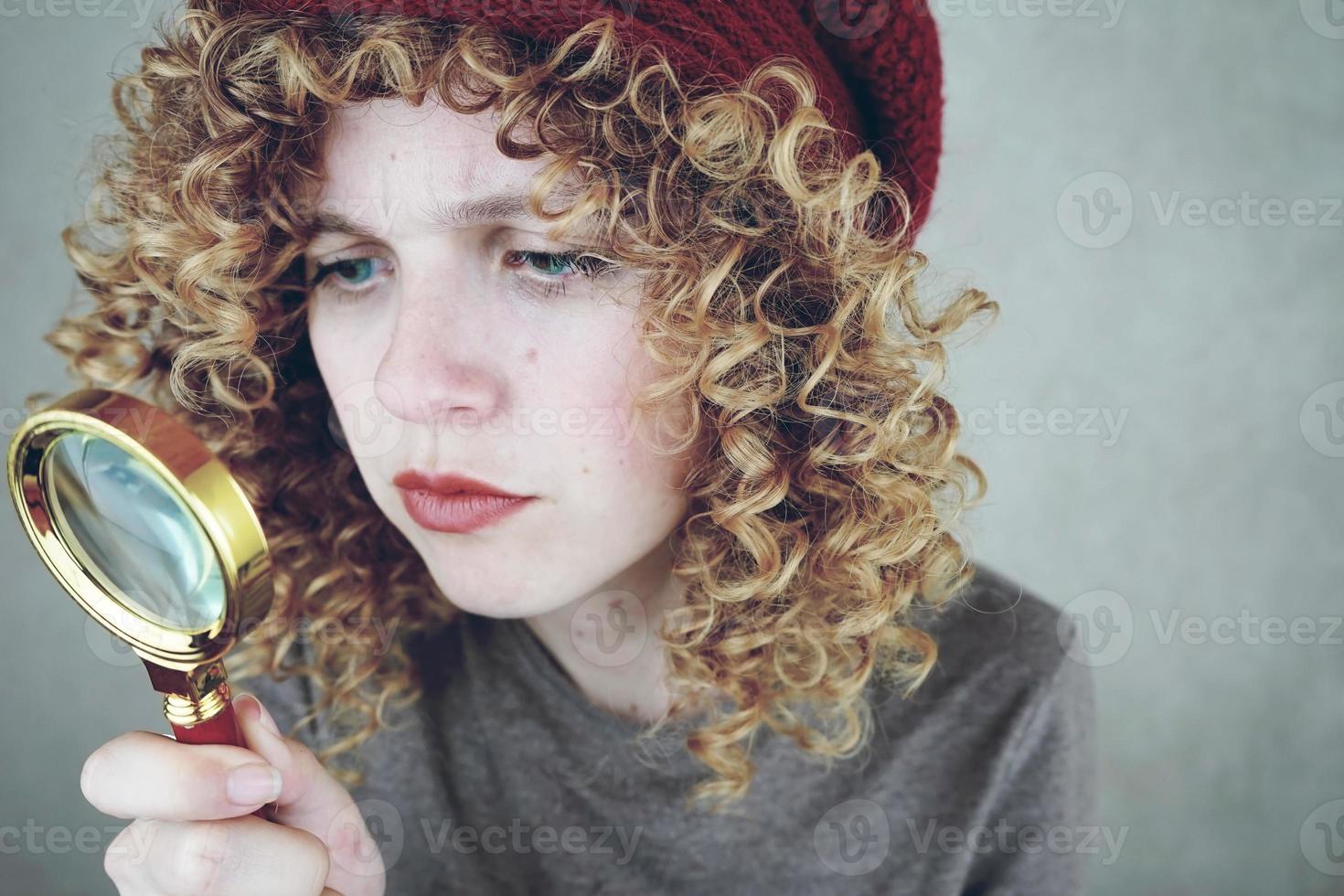 Closeup retrato de una bella y joven mujer divertida con ojos azules y cabello rubio rizado investigando con una lupa foto