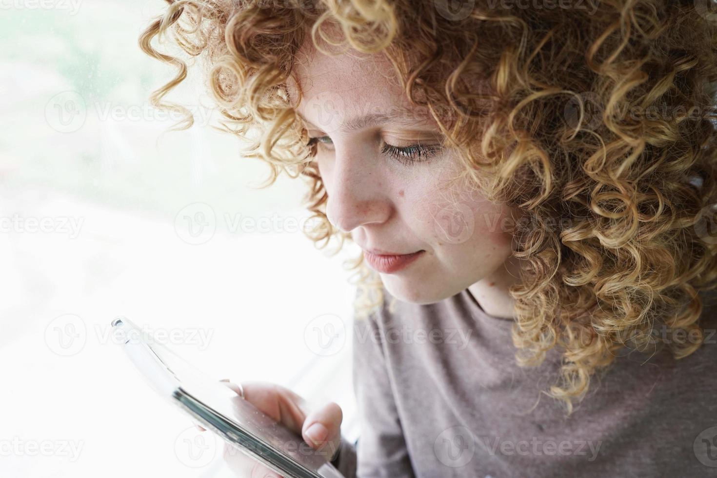 Closeup retrato de una bella y joven mujer con ojos azules y cabello rubio rizado mira el teléfono inteligente mientras ella está en un viaje foto